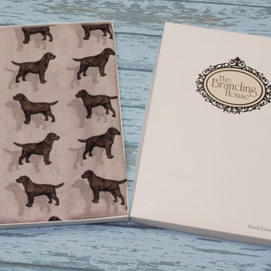 Può includere: Un tessuto marrone chiaro con un motivo ripetuto di cani Labrador marrone scuro, presentato in una scatola bianca aperta. Il coperchio della scatola mostra il testo The Branding House all'interno di una cornice decorata e Hand Printed In The UK.