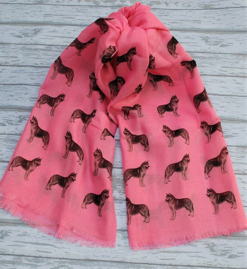 May include: A bright pink scarf featuring a repeating pattern of dark brown and black husky dog silhouettes. The lightweight fabric has a soft texture and finished edges with a delicate fringe detail along the bottom hem of the rectangular accessory.