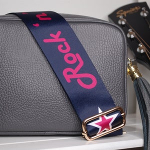 May include: A gray leather crossbody bag with a navy blue strap that says "Rock 'n' Roll" in pink lettering. The strap has a gold buckle with a red star. The bag has a gray tassel.