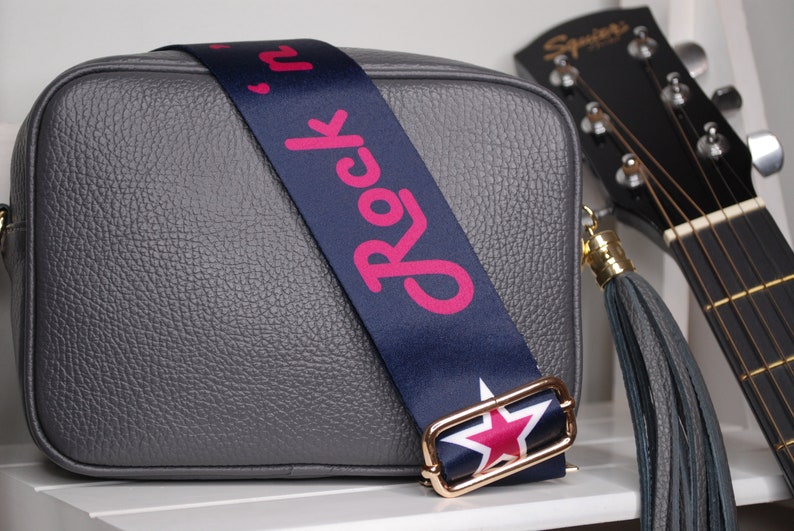 May include: A gray leather crossbody bag with a navy blue strap that says "Rock 'n' Roll" in pink lettering. The strap has a gold buckle with a red star. The bag has a gray tassel.