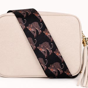 May include: A rectangular beige crossbody bag with a textured surface, featuring a wide black shoulder strap decorated with a repeating leopard print pattern. A long beige leather tassel hangs from the side, attached by gold-toned hardware.