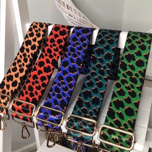 Puede incluir: Cinco correas ajustables para bolsos con un patrón repetido de leopardo negro sobre fondos naranja, rojo, azul, verde oscuro y verde brillante. Cada correa incluye herrajes de metal dorado, incluyendo deslizadores rectangulares y mosquetones giratorios.