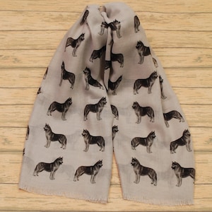 May include: A light beige scarf featuring a repeating pattern of dark gray and black husky dog illustrations. The scarf has a rectangular shape with frayed edges at the bottom, showcasing multiple dogs in various standing poses across the fabric surface.
