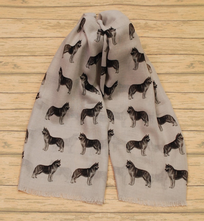 May include: A light beige scarf featuring a repeating pattern of dark gray and black husky dog illustrations. The scarf has a rectangular shape with frayed edges at the bottom, showcasing multiple dogs in various standing poses across the fabric surface.