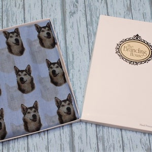 Pu&ograve; includere: Un tessuto azzurro con un motivo ripetuto di volti di cane husky, presentato in una scatola rettangolare aperta. Il coperchio della scatola &egrave; color crema con un logo nero incorniciato che recita The Branding House e Hand Printed In The UK.