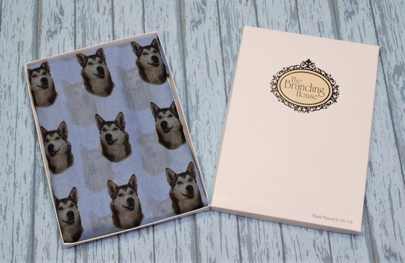 Pu&ograve; includere: Un tessuto azzurro con un motivo ripetuto di volti di cane husky, presentato in una scatola rettangolare aperta. Il coperchio della scatola &egrave; color crema con un logo nero incorniciato che recita The Branding House e Hand Printed In The UK.