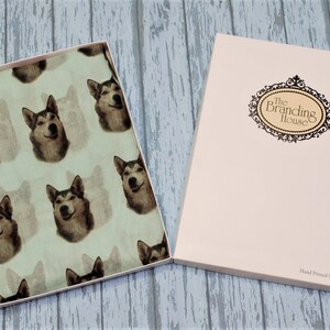 Pu&ograve; includere: Una scatola regalo rettangolare contenente un tessuto verde chiaro con un motivo ripetuto di volti di cane husky. Il coperchio bianco presenta una cornice ovale nera decorativa con il testo The Branding House e Hand Printed In The UK.