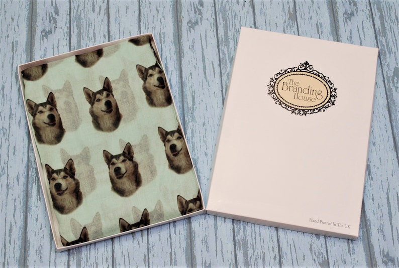Pu&ograve; includere: Una scatola regalo rettangolare contenente un tessuto verde chiaro con un motivo ripetuto di volti di cane husky. Il coperchio bianco presenta una cornice ovale nera decorativa con il testo The Branding House e Hand Printed In The UK.