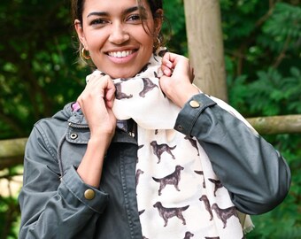 May include: A lightweight off-white scarf featuring a repeating pattern of dark brown Labrador retriever silhouettes. The scarf is worn draped around the neck, showcasing the detailed canine illustrations printed across the fabric surface.