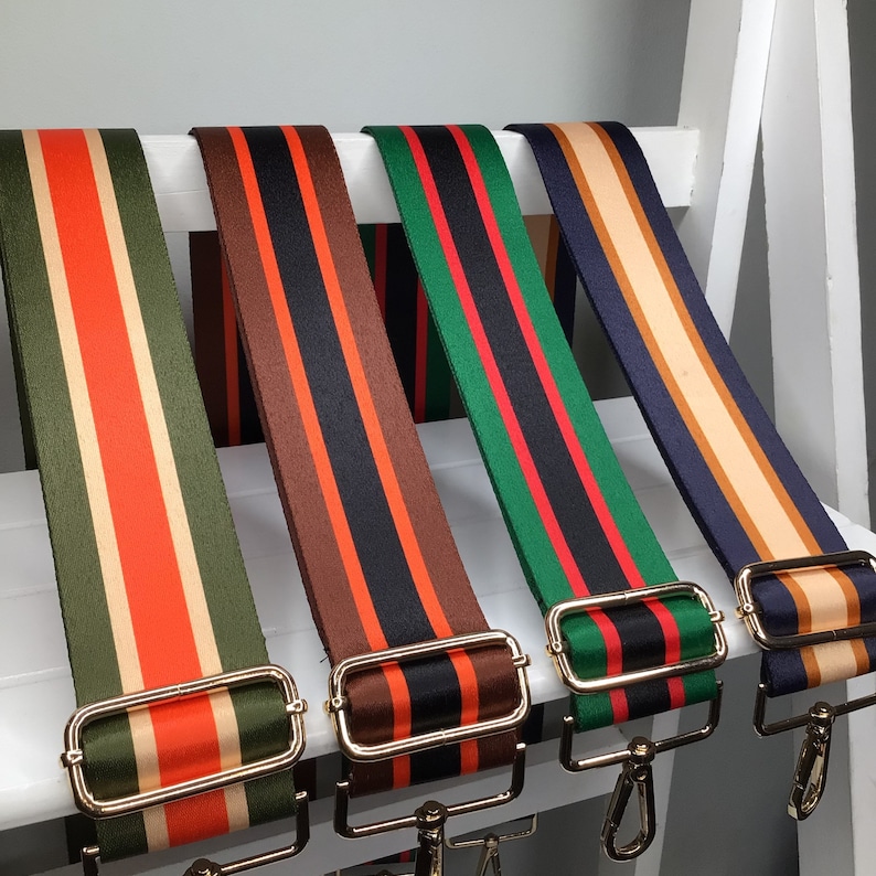 May include: Four colorful striped bag straps with gold-colored metal hardware. The straps feature vertical stripes in various color combinations: olive green, orange, and beige; brown, black, and orange; green, red, and black; and navy, beige, and brown.