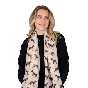 May include: A cream-coloured scarf featuring a repeating pattern of dark brown German Shorthaired Pointer dog silhouettes. The scarf is draped over a black jumper, showcasing the detailed canine print across the light fabric surface.