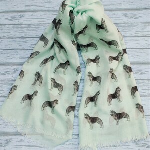 May include: A light mint green scarf featuring a repeating pattern of dark gray and white husky dogs in various standing poses. The rectangular scarf has a soft, lightweight appearance and is finished with delicate frayed fringe edges on both ends.