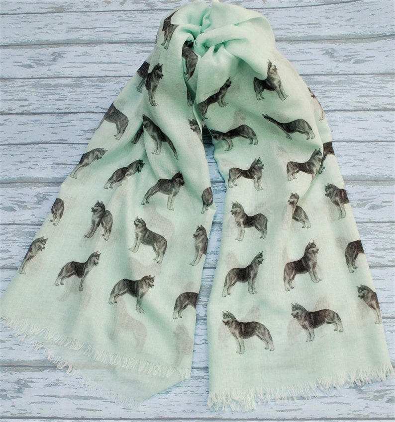 May include: A light mint green scarf featuring a repeating pattern of dark gray and white husky dogs in various standing poses. The rectangular scarf has a soft, lightweight appearance and is finished with delicate frayed fringe edges on both ends.