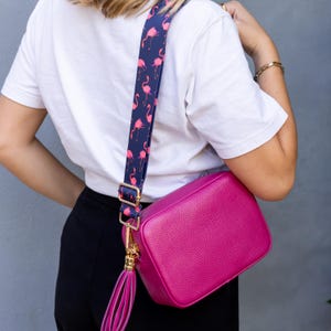 K&ouml;nnte beinhalten: Eine hellrosa Leder-Umh&auml;ngetasche mit goldenem Quastenanh&auml;nger und einem dunkelblauen und rosafarbenen Gurt mit Flamingomuster.