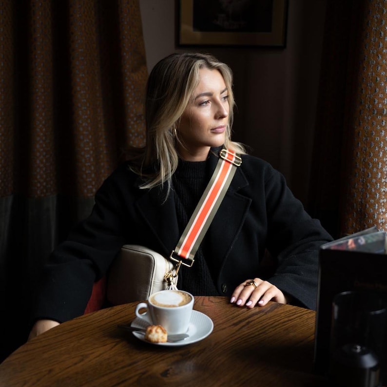 May include: A cream-colored crossbody bag with a striped strap in olive green, orange, and white. The bag is paired with a black coat and a cup of coffee on a wooden table. Gold hardware accents the strap.
