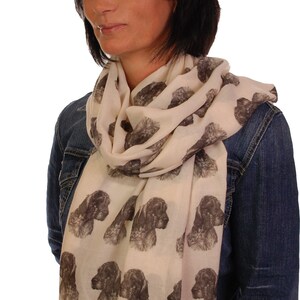 May include: A woman wearing a cream-colored scarf with a repeating pattern of brown and black dog heads. She is wearing a denim jacket and jeans.