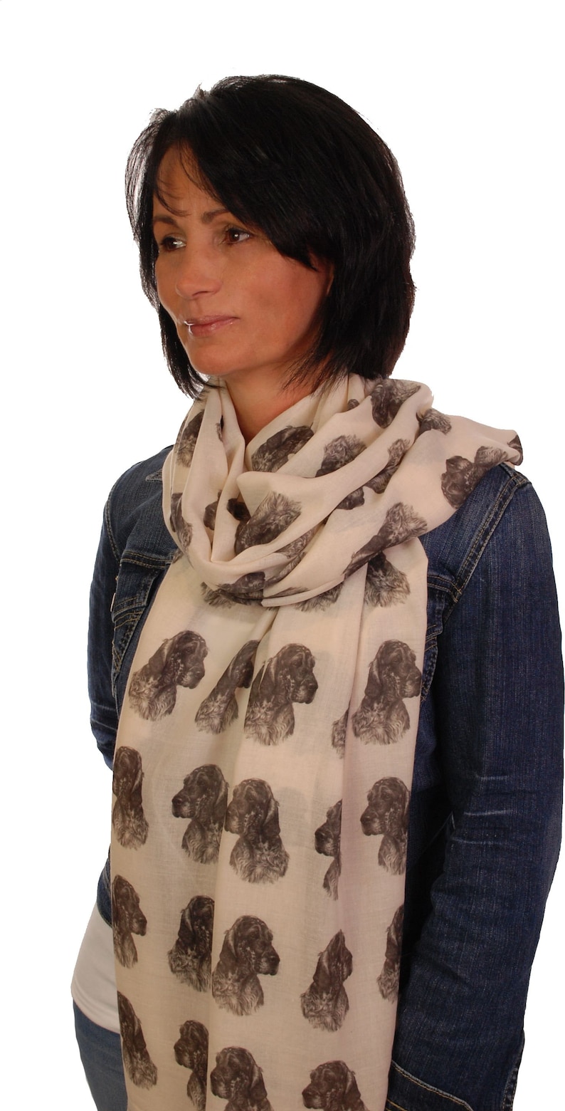 May include: A woman wearing a cream-colored scarf with a repeating pattern of brown and black dog heads. She is wearing a denim jacket and jeans.