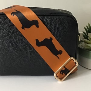 May include: A black leather crossbody bag with an orange strap featuring black dachshund silhouettes. The strap has a gold buckle.