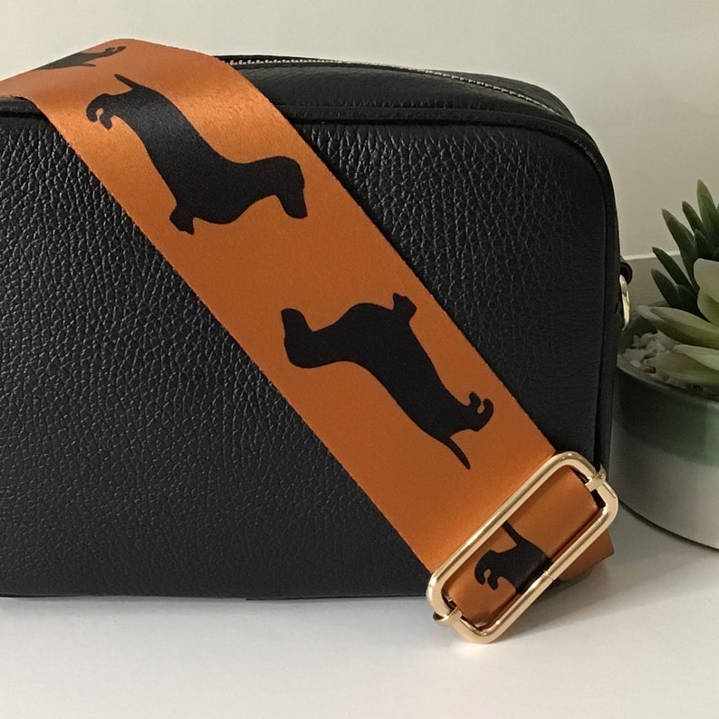 May include: A black leather crossbody bag with an orange strap featuring black dachshund silhouettes. The strap has a gold buckle.