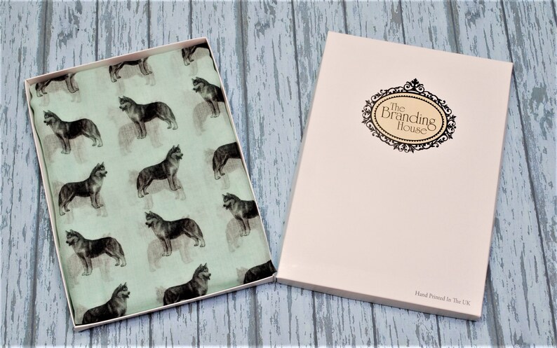 May include: A light green fabric scarf featuring a repeating pattern of dark gray husky dogs, presented in an open rectangular box. The box lid is cream-colored with a decorative black oval frame containing the text The Branding House.