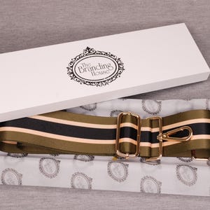 May include: A shoulder strap with olive green, black, and peach stripes, presented in a white box. The strap features gold-colored metal hardware and is packaged on patterned tissue paper. The box lid displays "The Branding House" logo.