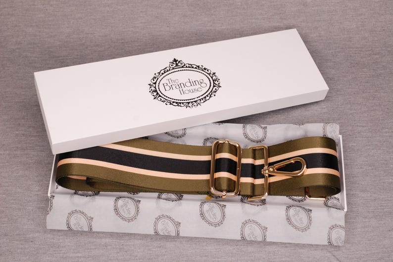 May include: A shoulder strap with olive green, black, and peach stripes, presented in a white box. The strap features gold-colored metal hardware and is packaged on patterned tissue paper. The box lid displays "The Branding House" logo.