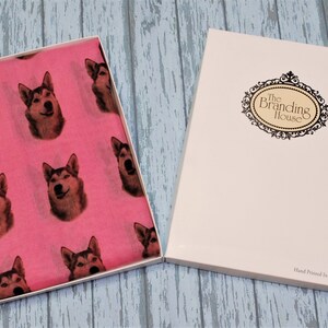 Pu&ograve; includere: Una sciarpa rosa brillante con un motivo ripetuto di volti di cane husky, presentata in una scatola regalo bianca aperta. Il coperchio della scatola mostra il testo The Branding House e Hand Printed In The UK in una cornice nera decorativa.