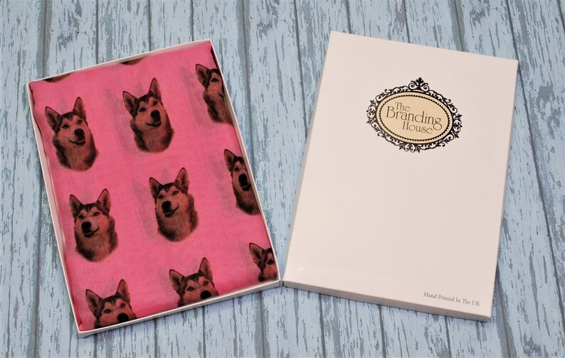 Pu&ograve; includere: Una sciarpa rosa brillante con un motivo ripetuto di volti di cane husky, presentata in una scatola regalo bianca aperta. Il coperchio della scatola mostra il testo The Branding House e Hand Printed In The UK in una cornice nera decorativa.