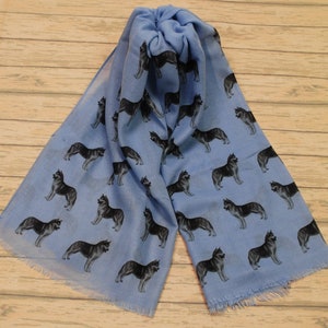 May include: A light blue scarf featuring a repeating pattern of black and grey husky dogs in various standing poses. The scarf has a soft, lightweight appearance with frayed edges at the bottom, showcasing a consistent print of the canine figures.