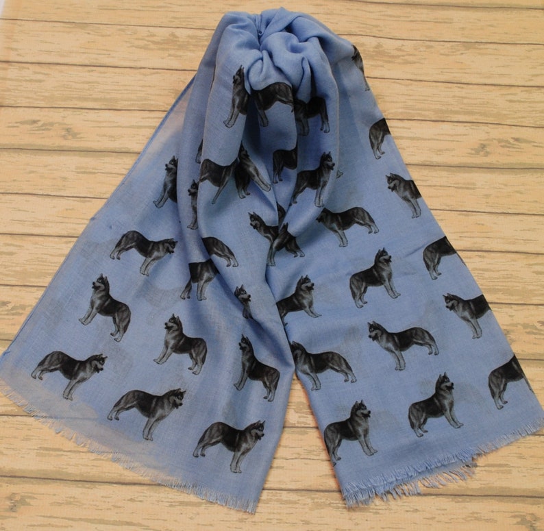 May include: A light blue scarf featuring a repeating pattern of black and grey husky dogs in various standing poses. The scarf has a soft, lightweight appearance with frayed edges at the bottom, showcasing a consistent print of the canine figures.