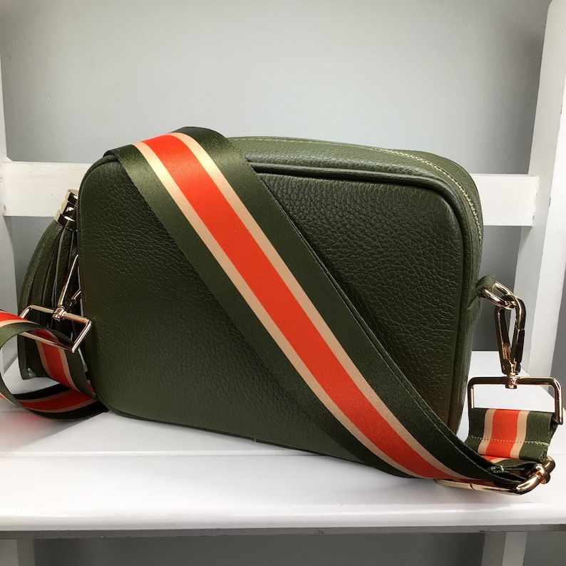 May include: Olive green crossbody bag with a textured finish. The bag features a wide strap with orange and beige stripes. Gold-colored hardware accents the bag's design, including the zipper and strap attachments.