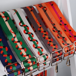 May include: A collection of colorful, patterned bag straps. Each strap features a repeating snake design in red, black, and white against a background of navy, green, white, brown, orange, and pink. The straps have gold-colored metal hardware.