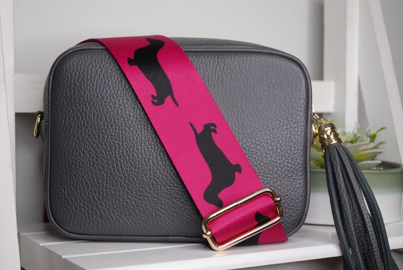 May include: A gray leather crossbody bag with a pink strap featuring black dachshund silhouettes. The bag has a gold buckle and a black tassel.