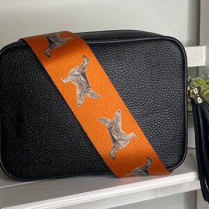 K&ouml;nnte beinhalten: Schwarze Leder-Kameratasche mit einem orangefarbenen Riemen mit einem sich wiederholenden Muster aus braunen Hunden. Die Tasche hat einen schwarzen Quasten.
