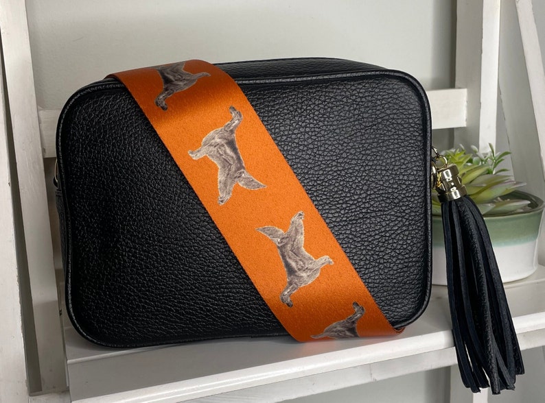 K&ouml;nnte beinhalten: Schwarze Leder-Kameratasche mit einem orangefarbenen Riemen mit einem sich wiederholenden Muster aus braunen Hunden. Die Tasche hat einen schwarzen Quasten.