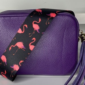 K&ouml;nnte beinhalten: Eine lilafarbene Leder-Umh&auml;ngetasche mit einem schwarzen und pinkfarbenen Flamingo-Muster-Riemen und einem lilafarbenen Quasten.