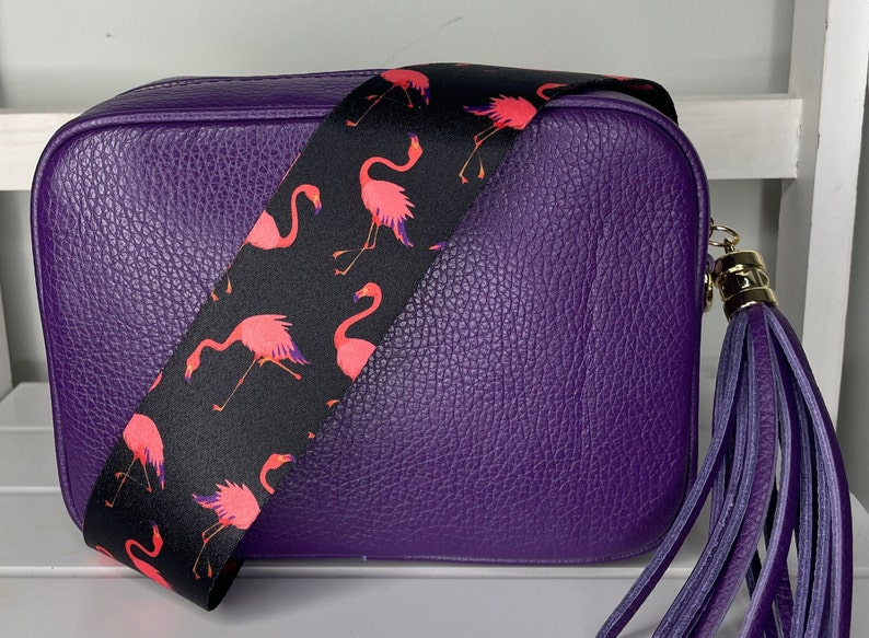 K&ouml;nnte beinhalten: Eine lilafarbene Leder-Umh&auml;ngetasche mit einem schwarzen und pinkfarbenen Flamingo-Muster-Riemen und einem lilafarbenen Quasten.