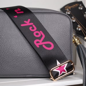 May include: A gray leather crossbody bag with a black strap that says "Rock 'n' Roll" in pink letters. The strap has a gold buckle and a pink star. The bag has a gray tassel.