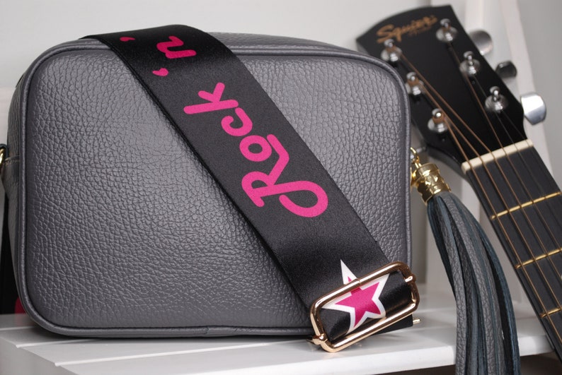 May include: A gray leather crossbody bag with a black strap that says "Rock 'n' Roll" in pink letters. The strap has a gold buckle and a pink star. The bag has a gray tassel.