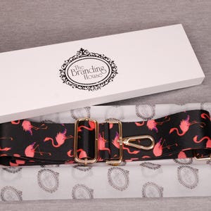 K&ouml;nnte beinhalten: Ein verstellbarer Gurt mit einem schwarzen und pinkfarbenen Flamingomuster und goldenen Beschl&auml;gen. Der Gurt ist in einer wei&szlig;en Box verpackt, auf der der Text "The Branding House" aufgedruckt ist.