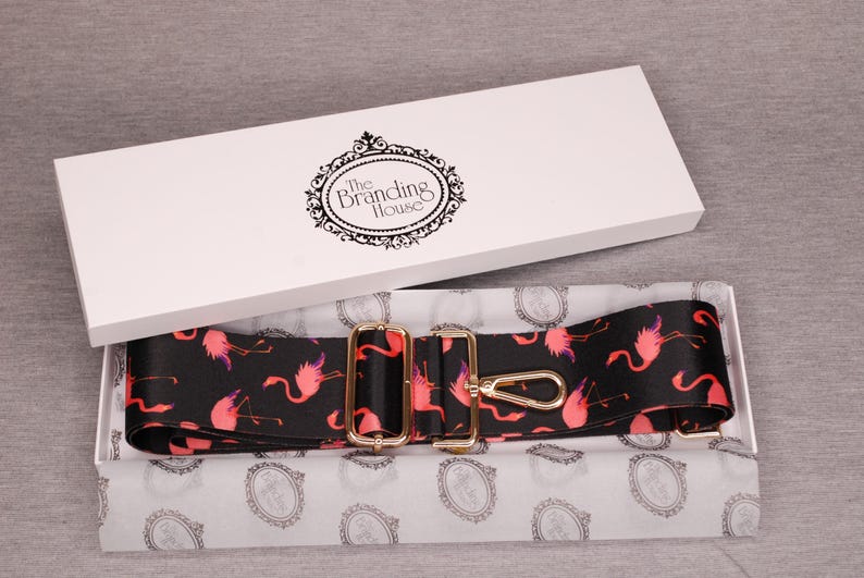 K&ouml;nnte beinhalten: Ein verstellbarer Gurt mit einem schwarzen und pinkfarbenen Flamingomuster und goldenen Beschl&auml;gen. Der Gurt ist in einer wei&szlig;en Box verpackt, auf der der Text "The Branding House" aufgedruckt ist.