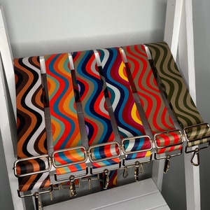 Puede incluir: Un conjunto de siete correas de colores con patrones ondulados y hebillas de metal dorado. Las correas están dispuestas en fila, con la correa más oscura a la izquierda y la más clara a la derecha.