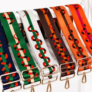 May include: Six adjustable bag straps featuring a repeating snake pattern in red, black, and white. The straps come in navy, green, white, brown, and two shades of orange, each equipped with gold-toned metal hardware, including sliders and swivel hooks.