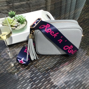 May include: A gray leather crossbody bag with a navy blue and white strap that says "Rock 'n' Roll" in pink lettering. The strap has a gold tassel.