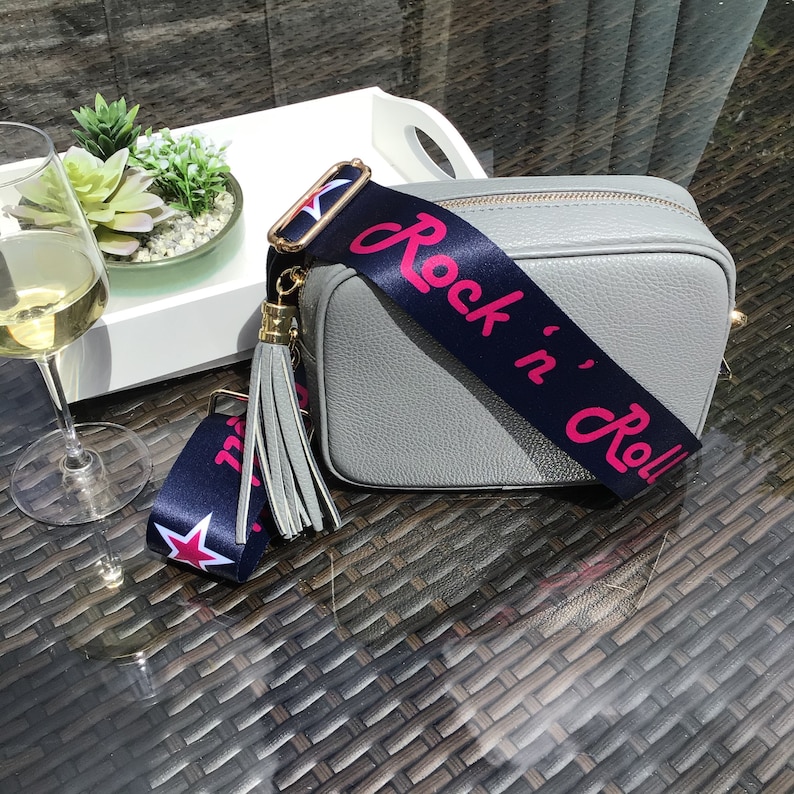May include: A gray leather crossbody bag with a navy blue and white strap that says "Rock 'n' Roll" in pink lettering. The strap has a gold tassel.