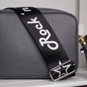 May include: A gray leather crossbody bag with a black adjustable strap that says "Rock 'n' Roll" in white lettering. The bag has a gold-colored zipper and a gray tassel.