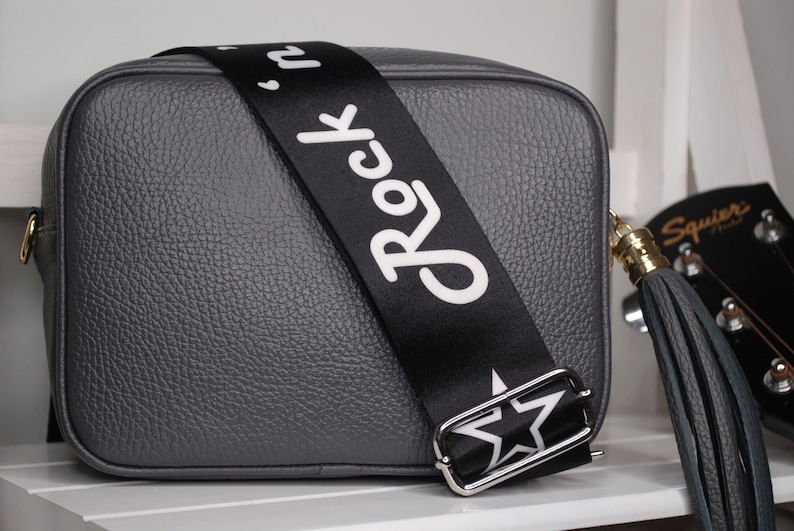 May include: A gray leather crossbody bag with a black adjustable strap that says "Rock 'n' Roll" in white lettering. The bag has a gold-colored zipper and a gray tassel.