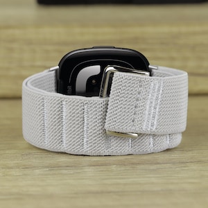 Könnte beinhalten: Ein graues elastisches Band mit einer silbernen Schnalle um eine schwarze Fitbit-Smartwatch.