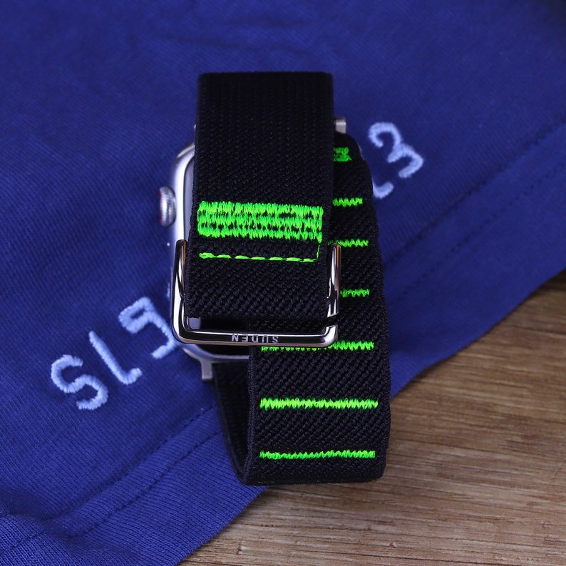 May include: A black and green watch band with the word "SLUDEN" printed on the metal buckle. The band has green stitching and is made of a woven material.