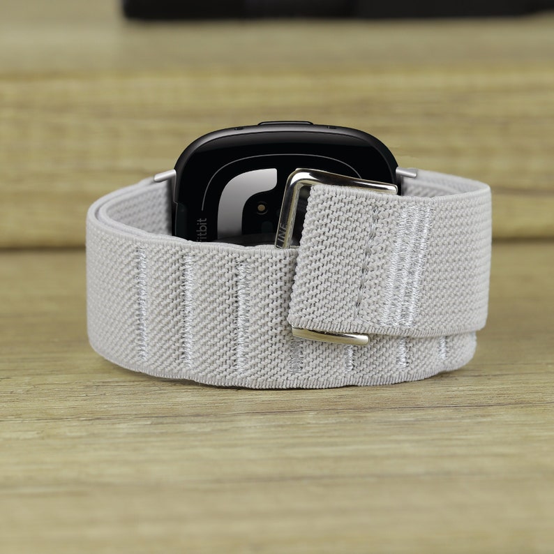 May include: A gray elastic band with a silver buckle wraps around a black Fitbit smartwatch.